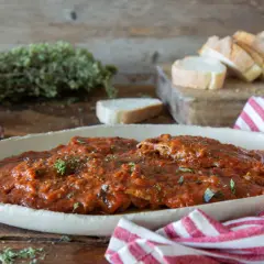 Herzhaftes Carne alla Pizzaiola in Tomatensauce, garniert mit Kräutern, neben Knoblauch, Brot und einem rot-weiß gestreiften Tuch.