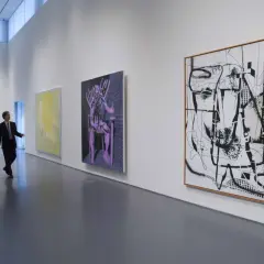 Un homme en costume observe une **analyse d'une œuvre d'art contemporain** dans une galerie. Des peintures abstraites et une sculpture colorée ornent les murs blancs.