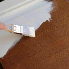 Mano pintando una puerta de madera exterior con brocha y pintura blanca.