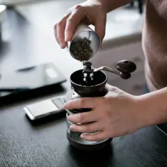Mani versano chicchi di caffè in un macinino manuale, iniziando la macinazione per un caffè perfetto.