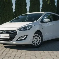Biały Hyundai i30 1.4 benzyna na tle zielonych drzew, zaparkowany na kostce brukowej.