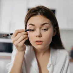 Dziewczyna wykonuje subtelny makijaż typu clean girl, precyzyjnie podkreślając oko kredką.