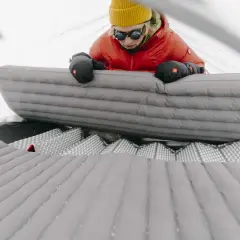 Osoba w pomarańczowej kurtce i żółtej czapce przygotowuje materac do spania w namiocie. Dba o wysoki r value, by zapewnić komfort w zimne dni.
