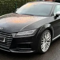 Negro Audi TT RS mojado por la lluvia, con reflejos naranjas en el capó. El coche está listo para la etapa 1 de su viaje.