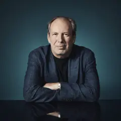 Hans Zimmer, der Meister der Filmmusik, sitzt mit verschränkten Armen vor einem dunklen Hintergrund. Sein Blick ist direkt auf die Kamera gerichtet.