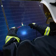 Strażak w białym hełmie i rękawicach zbliża iskrzący drut do paneli słonecznych, tworząc łuk elektryczny.