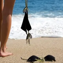 Nogi na plaży, obok leżą czarne bikini. Słońce grzeje, a fale obmywają brzeg. Czas na kąpiel dla golasy.