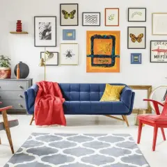 Salon w stylu eklektycznym: granatowa kanapa, czerwone fotele, galeria obrazów z motylami i owadami, szara komoda, dywan z geometrycznym wzorem.