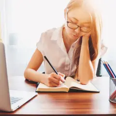 Dziewczyna z okularami pisze w zeszycie, ucząc się do egzaminu. To jak interaktywny kurs co to znaczy – nauka przez praktykę.