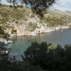 A serene view of Baia degli Infreschi, a secluded cove with clear turquoise waters, rocky cliffs, and lush greenery. A lone sailboat drifts peacefully in the bay.