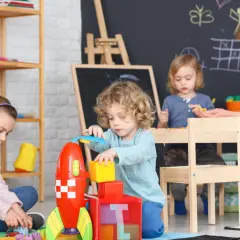 Dzieci bawią się klockami i rakietą. Czy praca w żłobku jest ciężka? Widok radosnych maluchów i opiekunki sugeruje, że niekoniecznie.