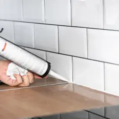 Application d'un joint d'étanchéité blanc sur un plan de travail en bois, près de carrelages muraux blancs. Le temps de séchage de la colle carrelage flex est une préoccupation pour une finition parfaite.
