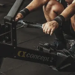 Mężczyzna ćwiczy na wioślarzu Concept 2. Pokazane są jego dłonie trzymające uchwyt i stopy na podnóżkach, co ilustruje, jak ćwiczyć na wioślarzu.