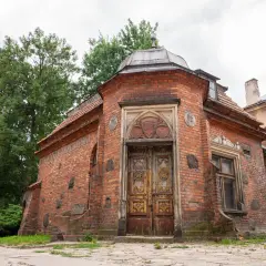 Stary ceglany budynek z ozdobnymi drzwiami i medalionami, otoczony zielenią.
