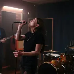 Wokalistka śpiewa do mikrofonu, basista gra na gitarze, a perkusista wybija rytm. Ich wspólna melodia wypełnia studio.