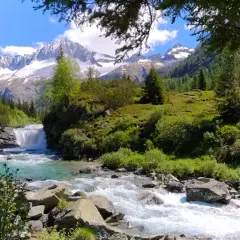 Un torrente impetuoso scorre tra rocce e vegetazione lussureggiante, con una cascata sullo sfondo. Le cime innevate delle montagne dominano il paesaggio del Val di Fumo, suggerendo l'alta altitudine.