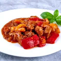 Gulasz z wołowiny z papryką, idealne mięso na gulasz. Aromatyczne danie na białym talerzu, ozdobione listkiem bazylii.
