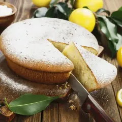 Una fetta di torta al limone morbidissima e profumatissima benedetta, cosparsa di zucchero a velo, con limoni freschi e foglie verdi su un tagliere di legno.