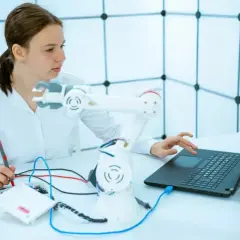 Junge Frau mit einem Roboterarm und Laptop, die an einem Projekt für ihr Robotik Studium arbeitet.