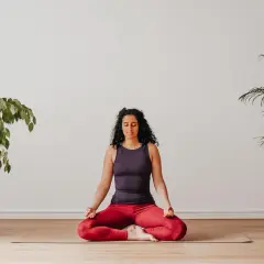 Frau praktiziert Yoga zu Hause im Lotussitz, umgeben von Pflanzen.