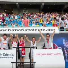 Zwei Kinder und zwei Erwachsene jubeln auf einer Tribüne vor einem Banner mit Bullen und einem Läufer.