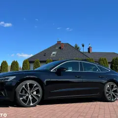 Elegancki czarny Audi A7 C8 lśni w słońcu na tle domu i zielonych drzew.