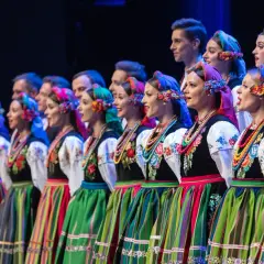 Zespół ludowy w tradycyjnych strojach, kobiety w kolorowych chustach i spódnicach, mężczyźni w tle.