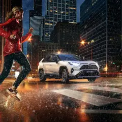 Kobieta biegnie w deszczu po mieście, obok niej jedzie biała Toyota RAV4 Hybrid. Zastanawiasz się, ile pali Toyota RAV4?