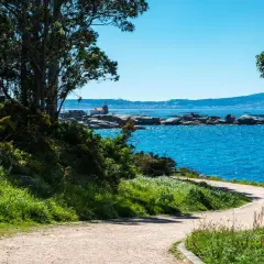 Sendero costero en la Illa de Arousa con mar azul brillante, rocas y un pequeño faro.