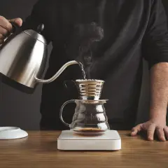 Mężczyzna przelewa gorącą wodę z czajniczka do drippera Kalita, przygotowując aromatyczną kawę.