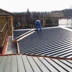 Operaio su tetto con muricci e tavelloni in fase di posa. Scaffolding e bandiera italiana sullo sfondo.