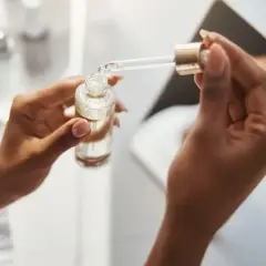 Dłonie trzymają buteleczkę z serum. Kropla z pipety spada do butelki, być może z kwasem ferulowym, dla zdrowej skóry.