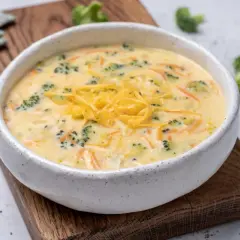 Aksamitna zupa brokułowa z serem cheddar, pełna zielonych różyczek i pomarańczowych nitek marchewki, podana w ceramicznej misce na drewnianej desce.