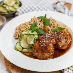 Två saftiga bitar sticky chicken med sesamfrön, serverade med nudlar och gurksallad.
