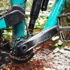 Close-up of a muddy mountain bike drivetrain with a teal frame, showing the crankset, chain, and pedal.
