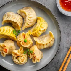Talerz pełen różnorodnych pierożków dim sum, z których każdy kusi innym kształtem i nadzieniem. Obok miseczka z ostrym sosem i pałeczki.