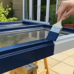 Main d'une personne peignant une fenêtre en bois déjà peinte avec une peinture bleu marine.
