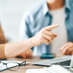 Dłoń wskazująca palcem na ekran laptopa, obok okulary i wykresy.