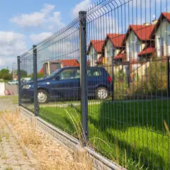 Płot z siatki z widocznym rozstawem słupków pod furtkę. W tle domy i samochody.