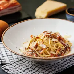 Un plato de spaghetti carbonara cremoso, con trozos de panceta crujiente y queso rallado. Ingredientes frescos como huevos, queso y pimienta negra completan la escena.