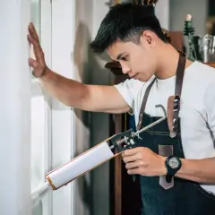 Un homme porte un tablier en jean et utilise un pistolet à calfeutrer pour recoller du verre sur un cadre de fenêtre blanc.