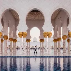 Un hombre camina bajo arcos blancos y dorados en Abu Dabi, reflejándose en el agua. Un visado Abu Dabi es el primer paso para disfrutar de esta maravilla.