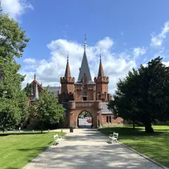Imponujący, czerwony zamek z wieżyczkami i bramą wjazdową, otoczony zielonym parkiem i drzewami.