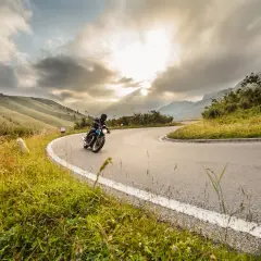 Un motociclista affronta una curva su una strada di montagna, godendosi un perfetto weekend in moto tra paesaggi mozzafiato e cielo nuvoloso.