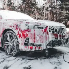 Czerwony samochód pokryty pianą, gotowy do mycia zimą. Wokół śnieg i piana, w tle drzewa.