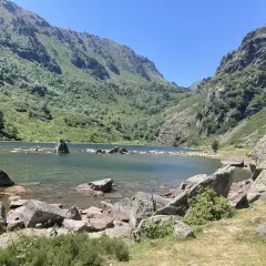 Lac de montagne entouré de montagnes verdoyantes et de rochers, sous un ciel bleu clair.