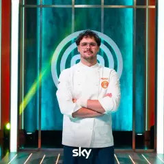 Matteo, vincitore MasterChef 15, posa con la sua divisa da chef, braccia conserte, sullo sfondo del logo del programma.