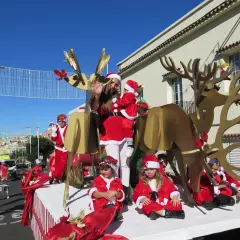 Niños y adultos disfrazados de Papá Noel y elfos participan en una cabalgata de Papá Noel, con un gran reno dorado y decoraciones navideñas.