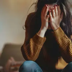 Kobieta w brązowym swetrze ukrywa twarz w dłoniach, wyrażając smutek. To może być obraz konsekwencji braku współżycia u kobiet.