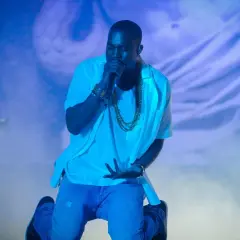 Kanye West na koncercie, klęczy z mikrofonem, otoczony niebieskim światłem i mgłą.